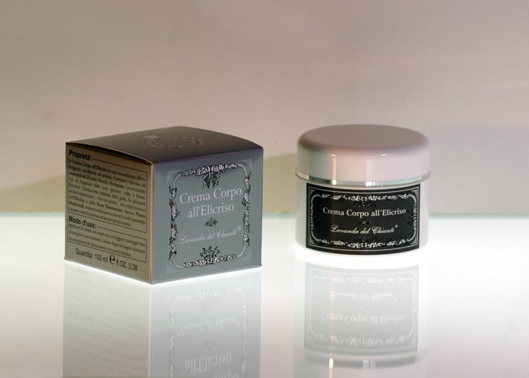 Body cream in a white plastic jar with silver line and silver box with white writing: "Helichrysum body cream". Bright light background with reflections on the table top
