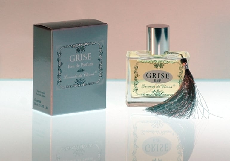 Flacone da profumo in vetro trasparente con guarnizione e tappo in argento con scritta Grise. Scatola argento con scritta: "Grise Eau de Parfume" di "Lavanda del Chianti". Sfondo luminoso chiaro con riflesso sul vetro