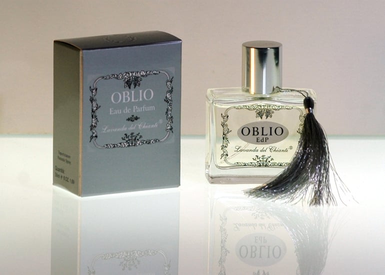 Flacone da profumo in vetro trasparente con guarnizione e tappo in argento con scritta Oblio. Scatola argento con scritta: "Oblio Eau de Parfume" di "Lavanda del Chianti". Sfondo luminoso chiaro con riflesso sul vetro