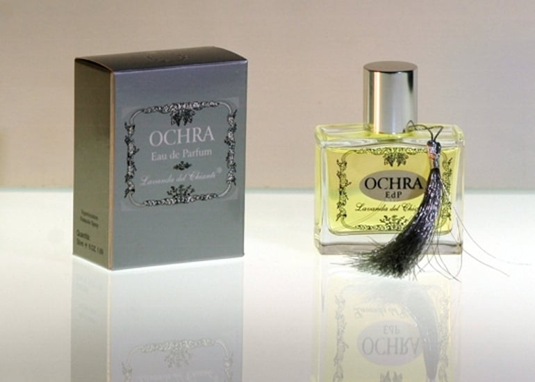 Perfume bottle in transparent glass with silver seal and cap with Ochra writing. Silver box with inscription: "Ochra Eau de Parfume" by "Lavanda del Chianti". Bright light background with reflection on the glass