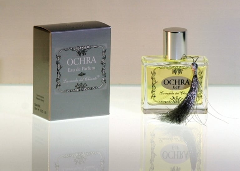 Flacone da profumo in vetro trasparente con guarnizione e tappo in argento con scritta Ochra. Scatola argento con scritta: "Ochra Eau de Parfume" di "Lavanda del Chianti". Sfondo luminoso chiaro con riflesso sul vetro