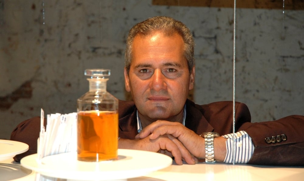 Doctor Lorenzo Domini with his medical experience applied to cosmetics and perfumery in amaranth jacket in front of a bottle of perfume on a rough masonry background