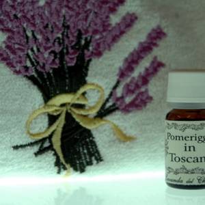 Su sfondo luminoso il particolare di una salvietta in cotone con mazzo di fiori di lavanda ricamato e piccolo flacone di profumo per ambiente con scritta: "Pomeriggio in Toscana"