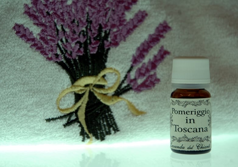 On a bright background, the detail of a cotton towel with an embroidered bunch of lavender flowers and a small bottle of home fragrance with the inscription: "Afternoon in Tuscany"
